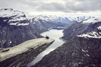 Paar sits on a rock platform on Trolltunga and looks over fjord landscape