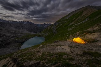 Tent in mountainous landscape with Lake Guffelsee in moonlight and cloudy sky