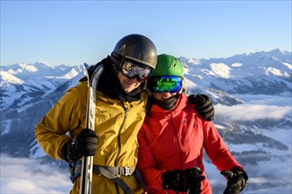 Two skiers with ski helmets and skis stand in front of a mountain panorama