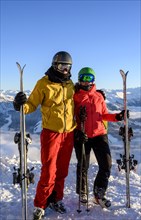 Two skiers with ski helmets and skis standing on the ski slope in front of a mountain panorama