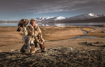 Mongolian eagle hunter posted with lake in front of landscape with lake and snow-covered mountains