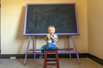 Friendly blonde boy sitting with a blackboard sponge by an empty blackboard