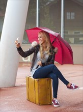 Girl with red umbrella sitting on a suitcase and making selfie, 16 years