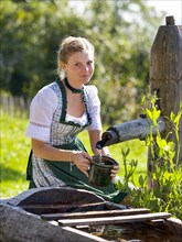 Young woman in dirndl fills a water jug at the well, 19 years