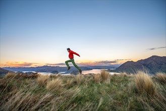 Hiker jumps into the air, panoramic view of Wanaka Lake and mountains at sunset