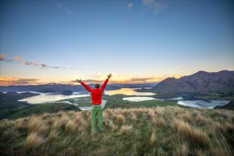 Hiker stretches his arms in the air, panoramic view of Wanaka Lake and mountains at sunset