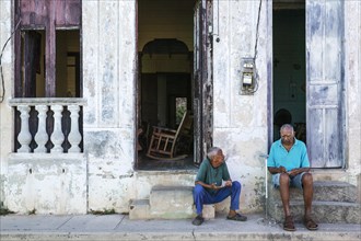 Two men having a chat at the doorsteps of their houses with decayed facades of crumbling plaster and weathered wooden doors