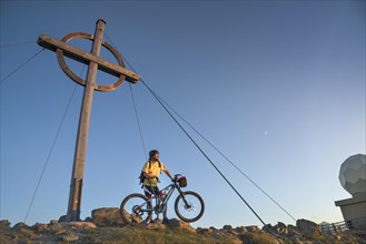 Mountain biker stands with e-MTB next to the summit cross of the Patscherkofel in the warm evening light