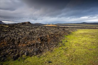 Black solidified lava borders on lush green grass with dramatic clouds in Leihrnjukur in Krafla