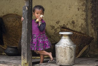A little girl stands in front of the house next to an aluminium water container and chews sugar cane