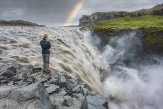 Blond woman with green down jacket standing at Dettifoss with rainbow