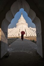 Buddhist monk stands with red umbrella in front of Hsinbyume Pagoda with frame of another pagoda