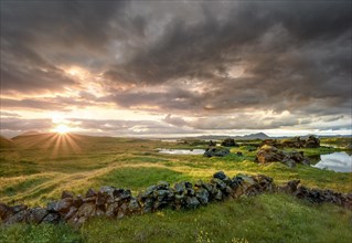 Stone wall and lava formations of volcanic rock rise from green meadows with dramatic sky and sun star in the evening light at Kalfastroend