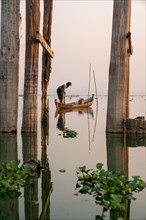Fisherman stands with boat on Taung Tha Man Lake for sunrise between the posts of the U-leg bridge