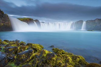 Long time exposure of a turquoise waterfall in volcanic landscape with dramatic clouds and green moss on rocks