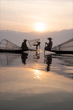 Three traditional fishermen pose standing and sitting on their small boats in front of the sun