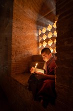 Buddhist young monk in red robe reading while sitting with a candle in front of rays of light in a temple