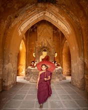 Buddhist young monk in red robe with red umbrella walks in a temple