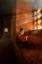 Buddhist young monk in red robe reading sitting in front of rays of light in a temple