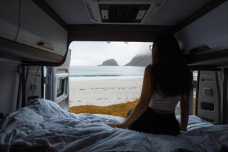 Frau sits on bed in campervan with view from rear of campervan on beach with turquoise water