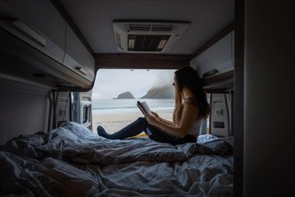 Frau reads on bed in campervan with view from rear of campervan on beach with turquoise water