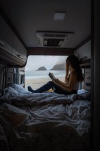 Woman reading on bed in campervan with view from rear of campervan on beach with turquoise water