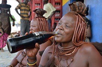 Traditionally dressed Himba woman drinking a coke in front of a bar, Opuwo, Kaokoland, Kunene, Namibia, Africa