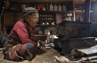An elderly woman sitting in front of a small stove in her kitchen