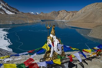 Tilicho Lake covered with ice in front of mountain scenery with Buddhist prayer flags and Hindu Shiva statue