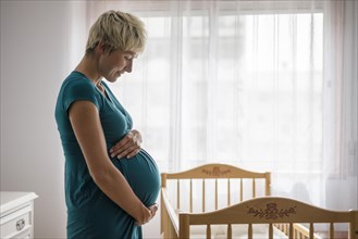 Pregnant woman holding her belly by baby cot next to the window
