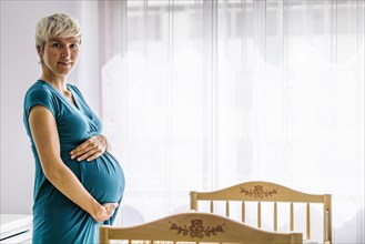 Pregnant woman holding her belly by baby cot next to the window