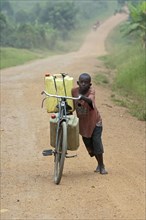 Boy pushing bicycle laden with full water containers along dusty road