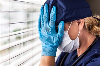 Female doctor or nurse with head in hands at window wearing medical face mask and surgical gloves