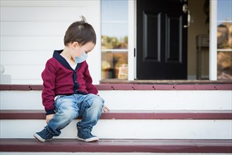 Melancholy lonely mixed race boy on front porch wearing medical face mask during the coronavirus pandemic