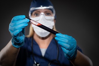 Female Lab Worker Wearing Medical Face Mask Holds Test Tube of Blood Against Dark Background