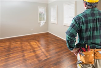 Contractor wearing toolbelt and hard hat facing empty room with hard wood floors
