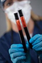 Female Lab Worker Wearing Medical Face Mask Holds Test Tubes of Blood Against Dark Background