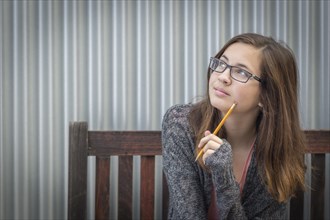 Pretty young daydreaming female student with pencil sitting on bench looking to the side