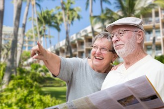 Happy senior adult couple tourists with brochure next to tropical hotel and palm trees
