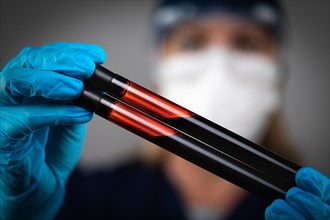 Female Lab Worker Wearing Medical Face Mask Holds Test Tubes of Blood Against Dark Background