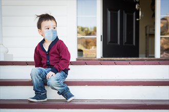 Melancholy lonely mixed race boy on front porch wearing medical face mask during the coronavirus pandemic