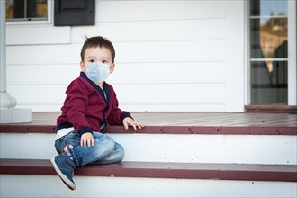 Melancholy lonely mixed race boy on front porch wearing medical face mask during the coronavirus pandemic