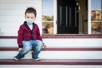 Melancholy lonely mixed race boy on front porch wearing medical face mask during the coronavirus pandemic