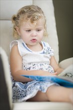 Adorable blonde haired blue eyed little girl reading her book in the chair