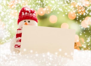Cute snowman with scarf and hat next to blank white card over abstract snow and light background