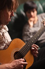 Young musician plays his acoustic guitar as friend in the background listens