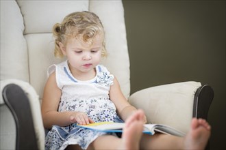 Adorable blonde haired blue eyed little girl reading her book in the chair