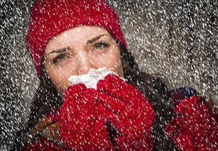 Sick mixed-race woman wearing winter hat and gloves blowing her sore nose with a tissue in the snow