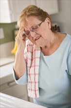 Grimacing senior adult woman at kitchen sink with head ache