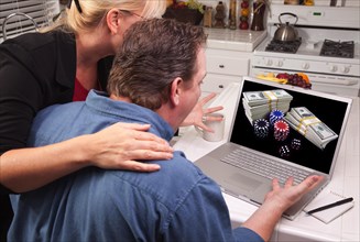 Couple in kitchen using laptop with stacks of money and poker chips on the screen